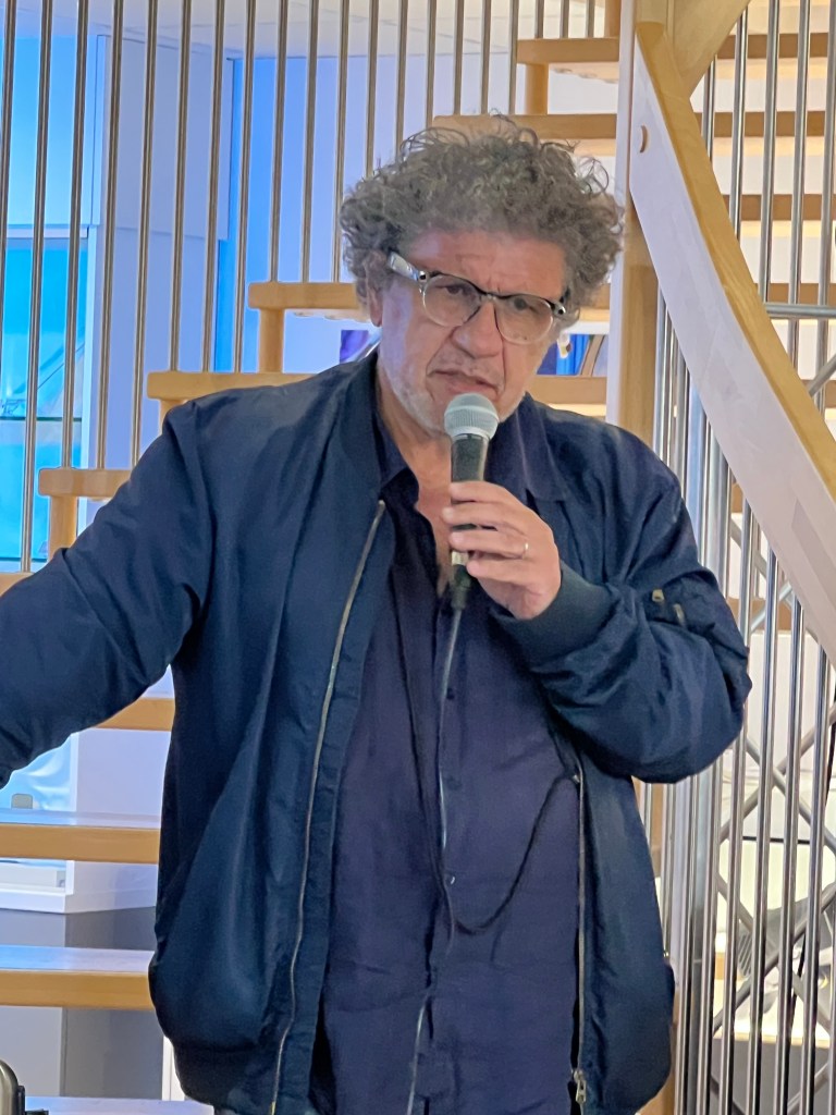 Un homme avec des cheveux bouclés et des lunettes, tenant un microphone sur un podium près d'un escalier en bois.