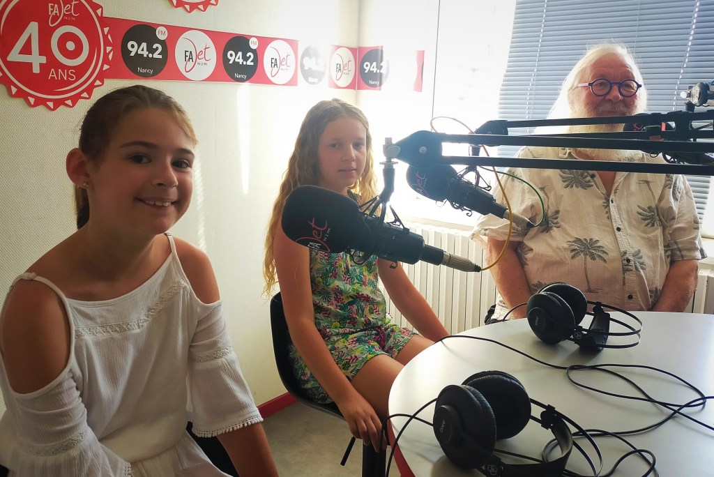 Deux jeunes filles et un homme âgé assis autour d'une table de studio de radio, avec des casques et des microphones devant eux, dans un environnement lumineux.