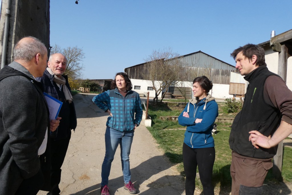 Un groupe de personnes discutant ensemble sur une exploitation agricole en plein air, avec des bâtiments agricoles en arrière-plan.
