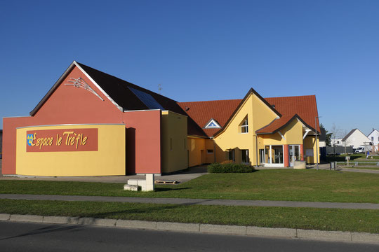 Vue extérieure d'un bâtiment coloré, l'Espace le Trèfle, avec un toit en tuiles rouges et des murs jaunes et terracotta, situé dans un espace vert.