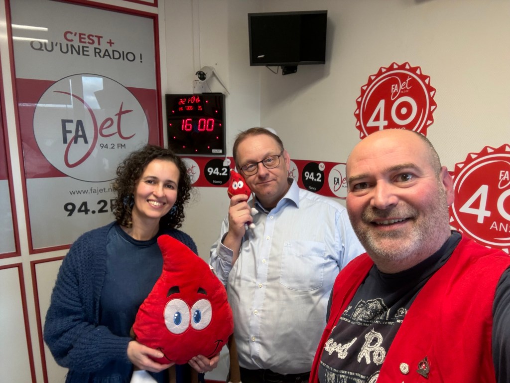 Trois personnes posant dans un studio de radio, entourées d'affiches et d'une horloge indiquant 16h00. Une personne tient un coussin en forme de goutte d'eau souriante.