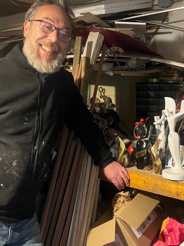 Un homme souriant avec une barbe, portant une veste noire, se tenant à côté d'un établi rempli d'objets d'art et de matériel, dans un atelier en désordre.