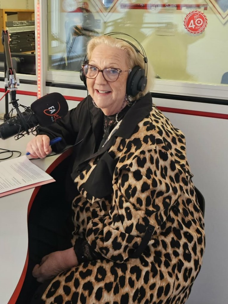 Femme souriante portant une veste en léopard et des écouteurs, assise devant un microphone dans un studio de radio.