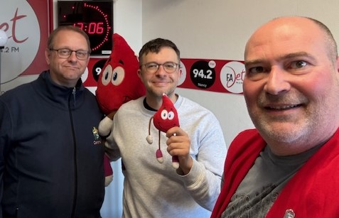 Trois hommes posant ensemble dans un studio de radio, tenant des peluches rouges, avec une horloge et un logo de radio en arrière-plan.