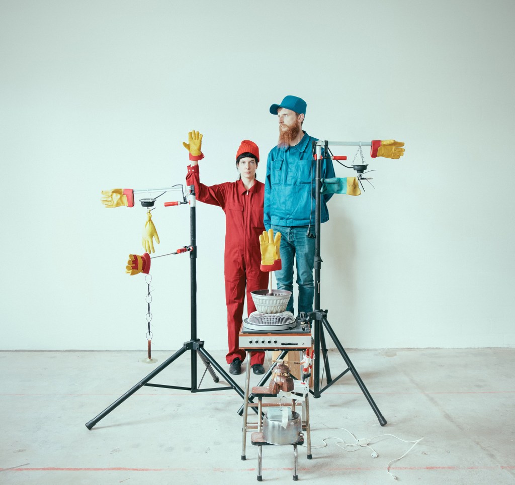 Deux personnes, l'une en salopette rouge et l'autre en tenue bleue, posent avec des gants en caoutchouc sur des supports en métal qui tiennent des objets ménagers.