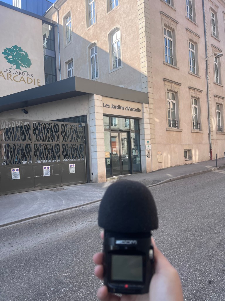 Une personne tient un enregistreur audio devant l'entrée de l'établissement Les Jardins d'Arcadie.