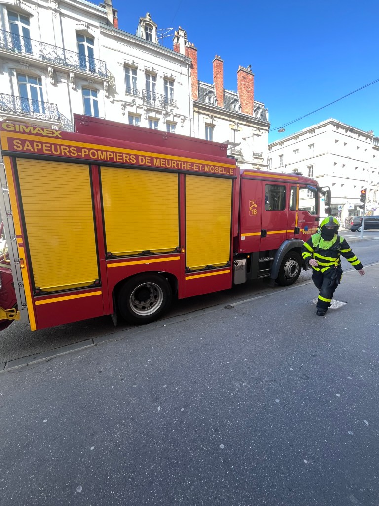 Camion de pompier rouge avec des détails jaunes, stationné sur une rue, un sapeur-pompier en uniforme s'approche.