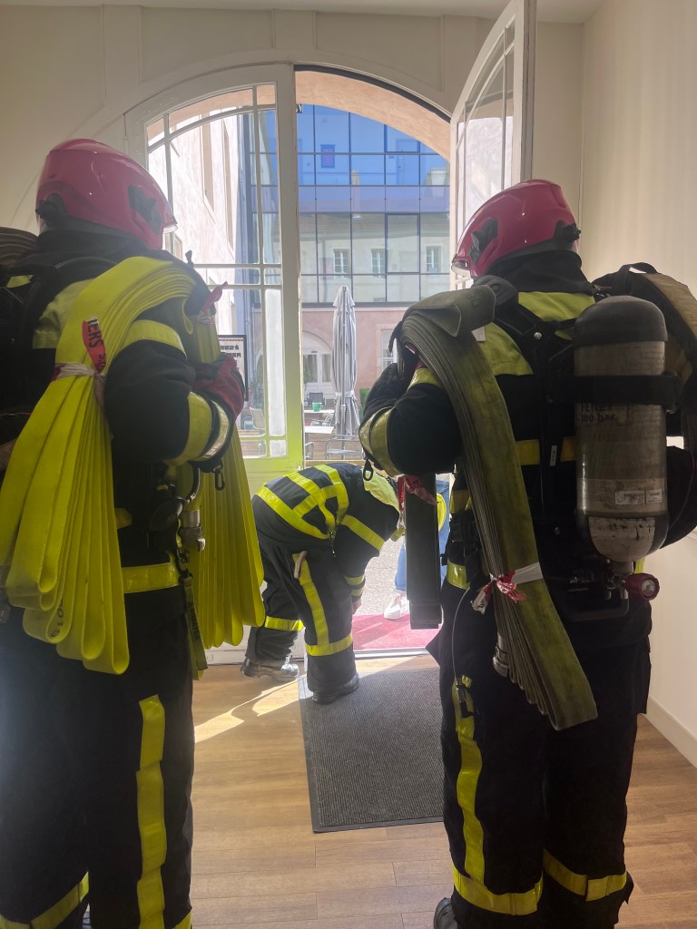 Trois pompiers en uniforme s'apprêtent à entrer dans un bâtiment, avec des équipements de lutte contre les incendies et des tuyaux dans les mains.