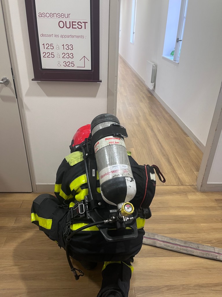 Un pompier équipé d'un appareil respiratoire s'agenouille dans un couloir devant un panneau indiquant l'ascenseur OUEST et les numéros d'appartements.