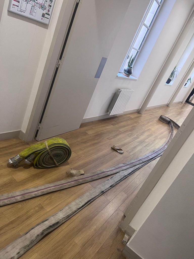 Un couloir avec un tuyau d'incendie enroulé et étendu sur le sol, à côté d'une porte fermée et de fenêtres laissant passer la lumière.