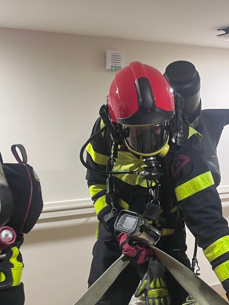 Un pompier en tenue de protection avec un casque rouge, utilisant un tuyau dans un couloir.