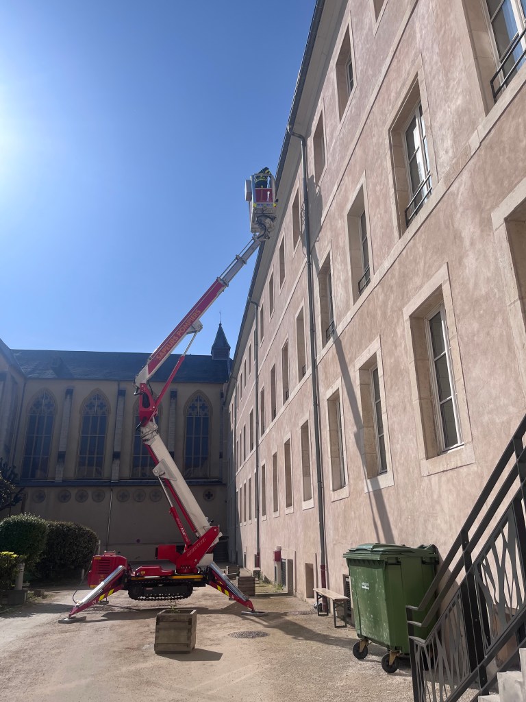 Un élévateur en activité à proximité d'un bâtiment, avec des travailleurs au sommet, sous un ciel bleu.