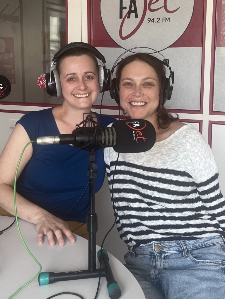 Deux femmes souriantes avec des casques audio, posant devant un micro dans un studio de radio.