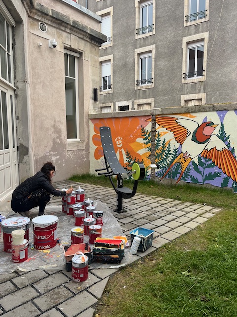 Une artiste peint un mur coloré avec un oiseau et des fleurs, entourée de pots de peinture sur un sol en pavés.