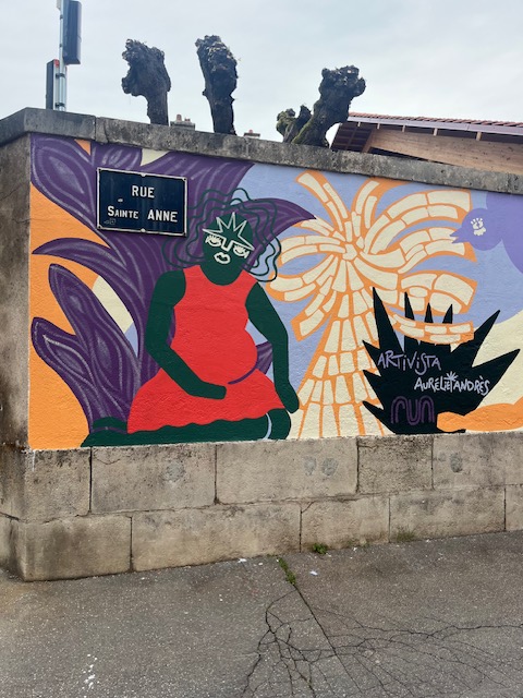 Mural coloré sur un mur montrant une femme avec des lunettes, assise au milieu de motifs floraux, portant un haut rouge et un bas vert. La rue Sainte Anne est indiquée sur un panneau.