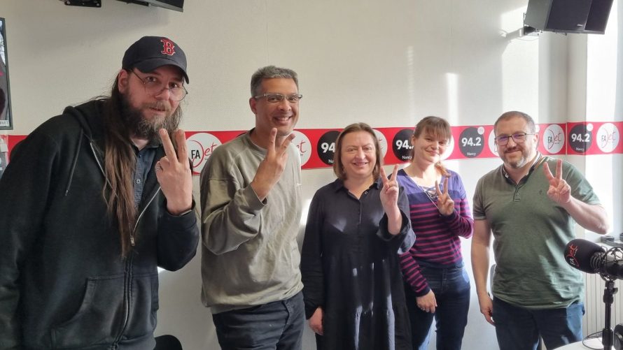 Cinq personnes faisant un signe de paix avec les mains, dans un studio de radio, avec des éléments de décor aux couleurs vives en arrière-plan.