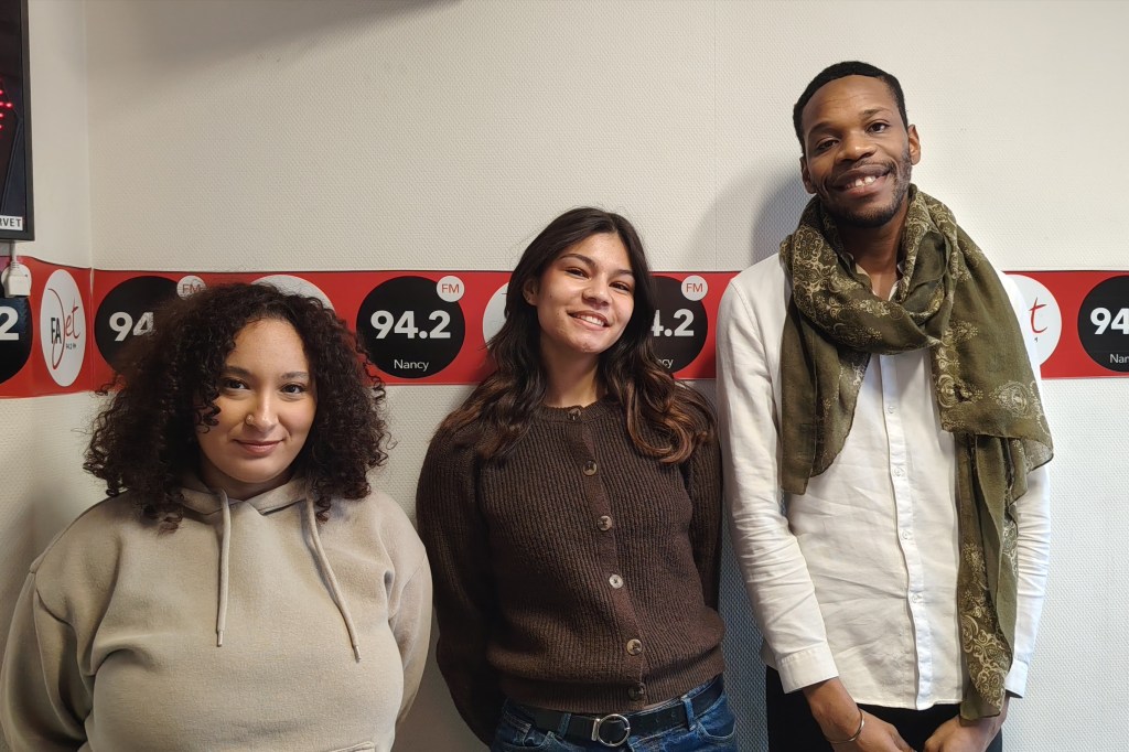Trois personnes posent ensemble dans un studio de radio, avec des panneaux affichant '94.2 FM Nancy' en arrière-plan.