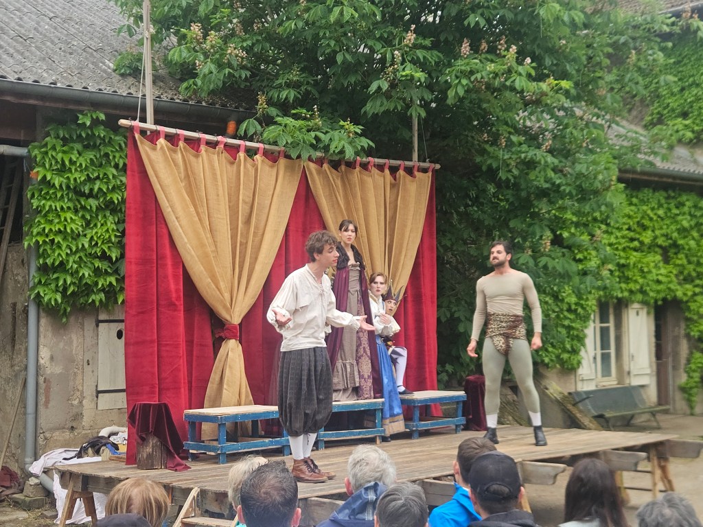 Représentation théâtrale en plein air avec des acteurs habillés en costumes historiques sur scène, devant un public attentif.