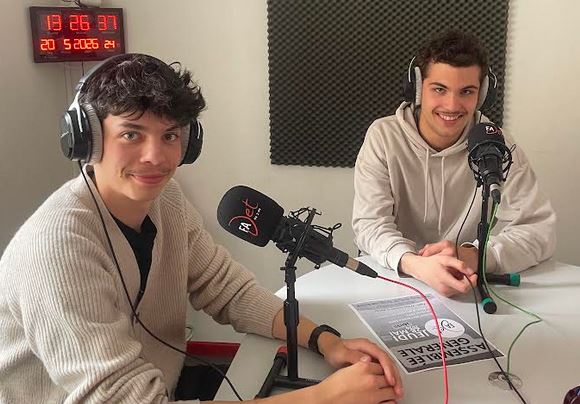 Deux jeunes hommes souriants portant des écouteurs, assis à une table d'enregistrement en studio de radio, avec un microphone et un panneau d'affichage en arrière-plan.