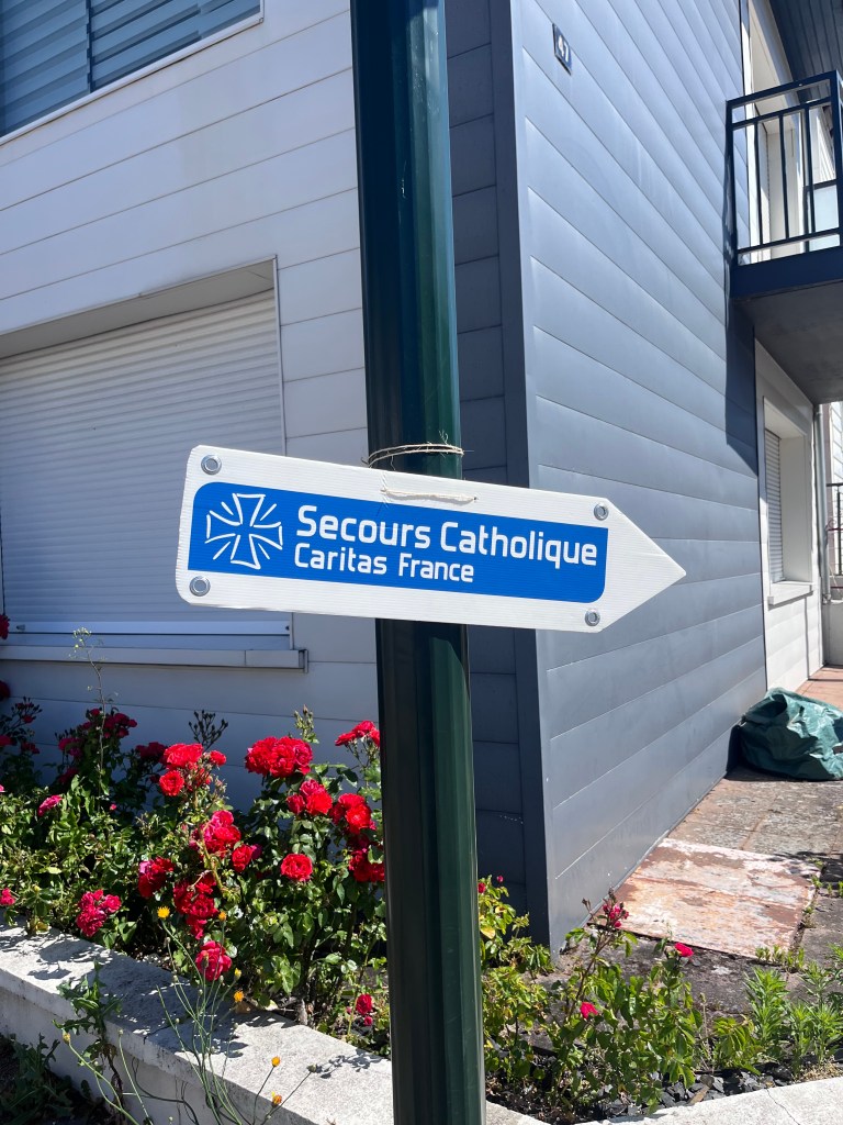 Panneau directionnel indiquant 'Secours Catholique Caritas France' avec des fleurs rouges en arrière-plan.