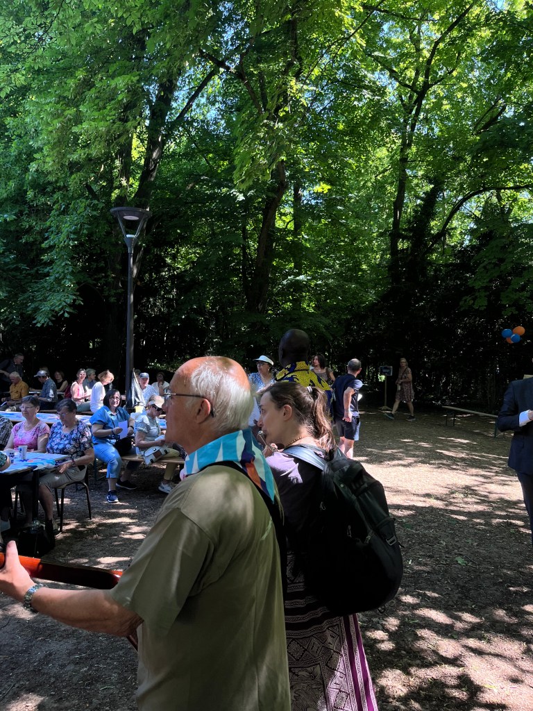 Une scène de rassemblement en plein air, avec des participants aux alentours d'une table, sous des arbres verts, tandis qu'un homme joue de la guitare.