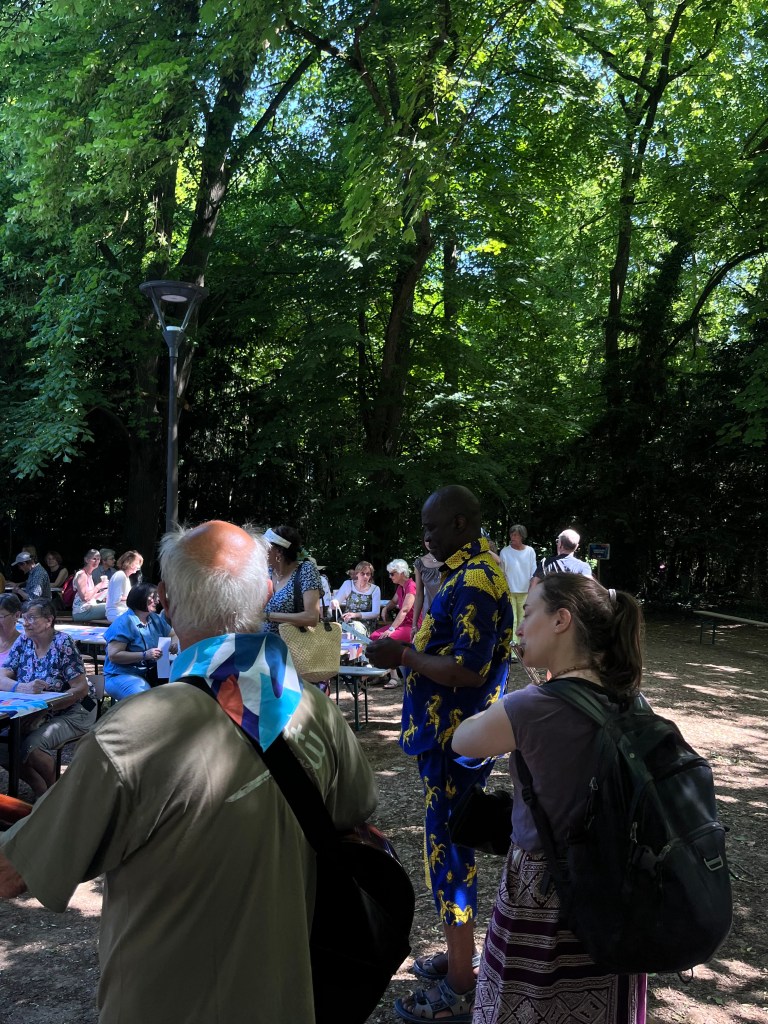 Groupe de personnes dans un parc, entouré d'arbres verdoyants, interagissant autour de tables lors d'un événement en plein air.
