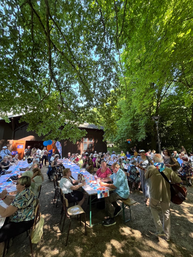 Un grand rassemblement de personnes assises autour de longues tables à l'extérieur, sous des arbres, lors d'un événement festif.