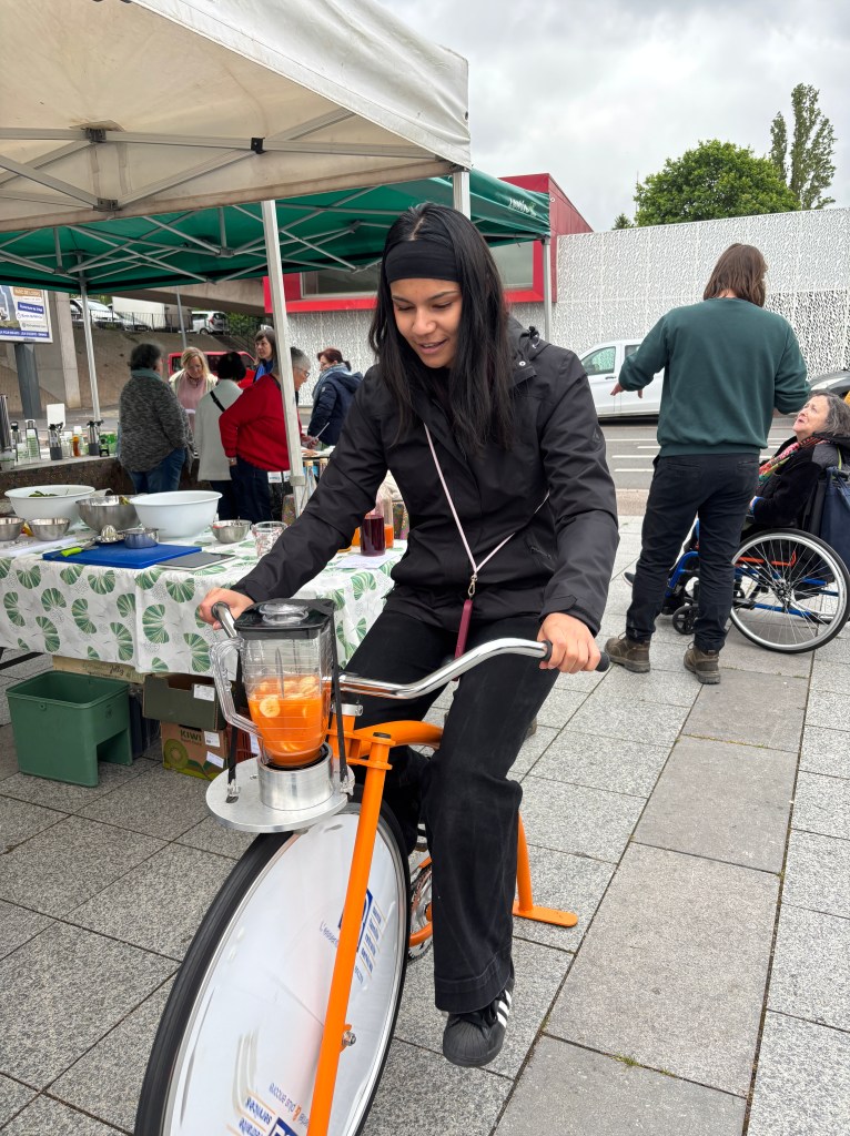 Une femme utilisant un vélo équipé d'un mixeur pendant un événement extérieur, avec des gens en arrière-plan.