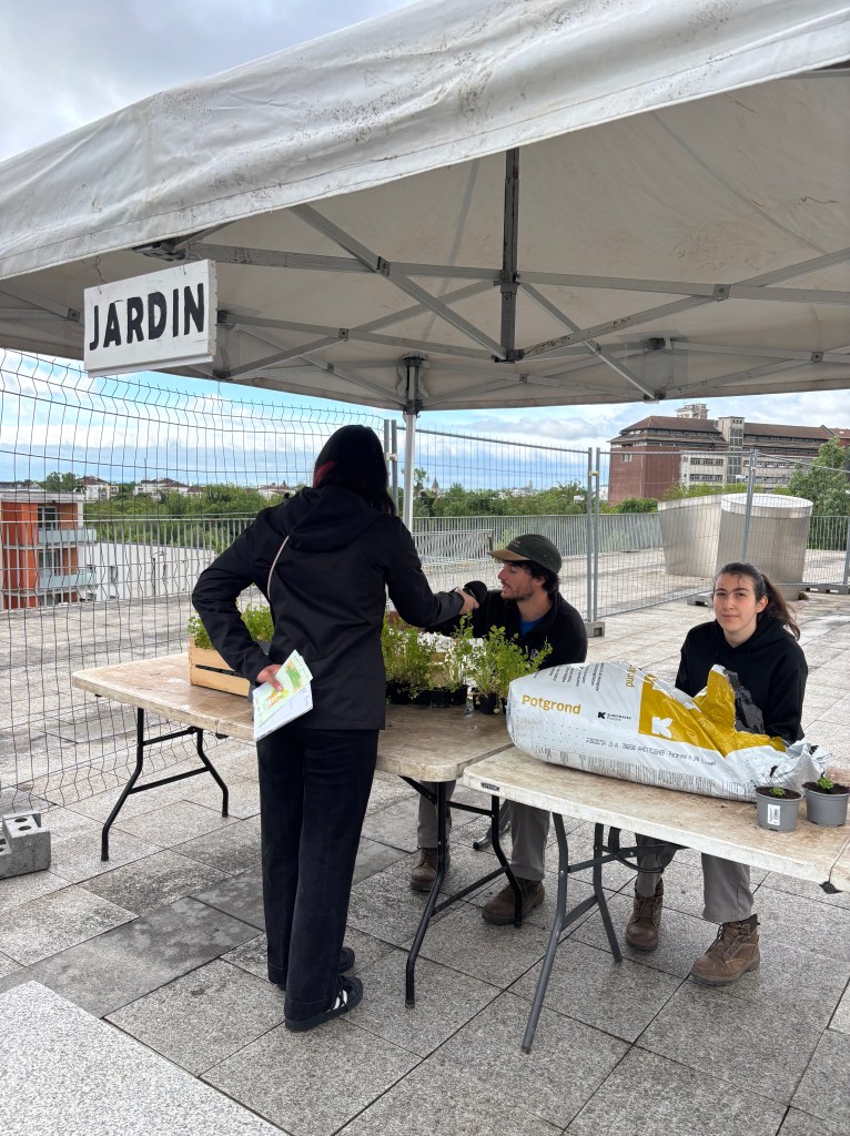 Un stand de jardinage sous un abri avec une enseigne 'JARDIN'. Des personnes discutent et partagent des plantes en pot.