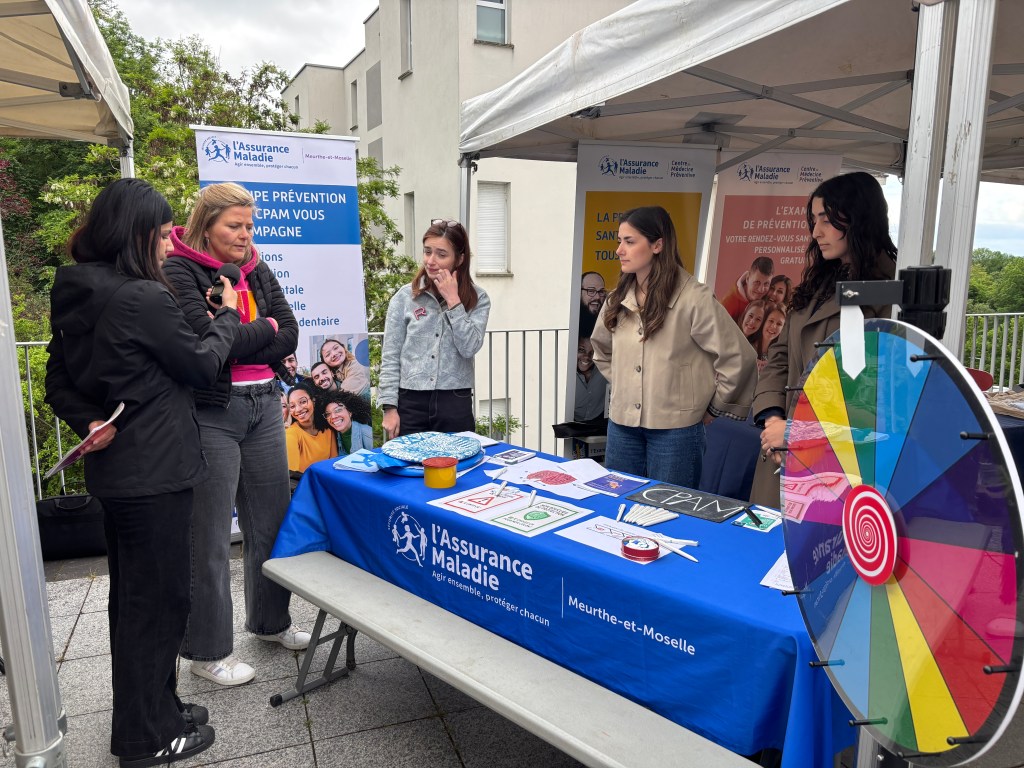 Un groupe de femmes discutant devant un stand de sensibilisation de l'Assurance Maladie, avec des affiches et des jeux éducatifs sur la santé.