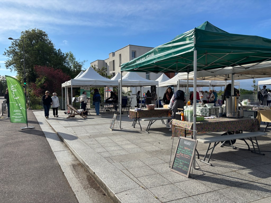 Marché en plein air avec des stands sous des tentes, des visiteurs déambulant entre les tables, et une signalétique verte.
