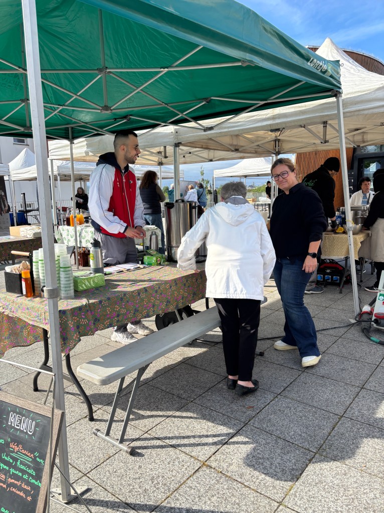 Un stand de marché extérieur avec un homme souriant en t-shirt rouge servant des boissons à une femme âgée en veste blanche, tandis qu'une autre femme les regarde. Des tentes blanches et un ciel bleu en arrière-plan.