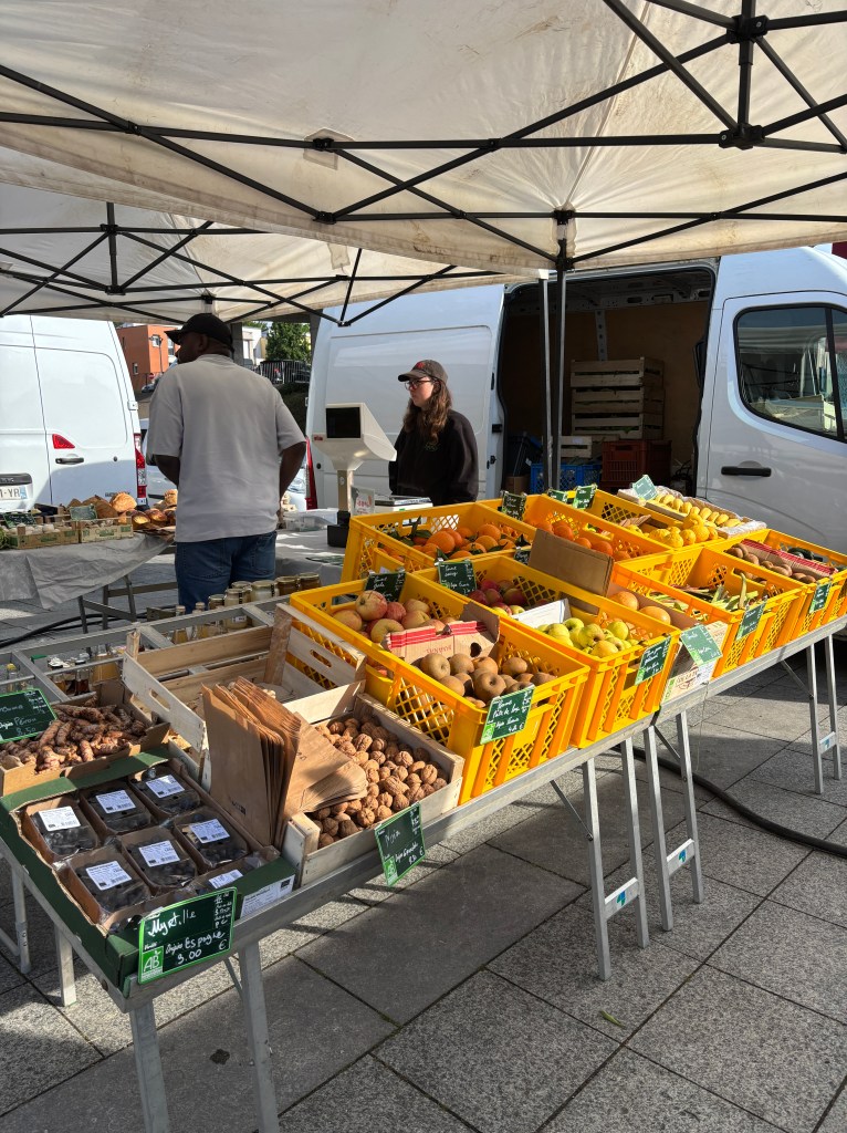 Un marché en plein air avec des étals de fruits et légumes, des caisses jaunes remplies de produits frais, et deux vendeurs derrière le comptoir.