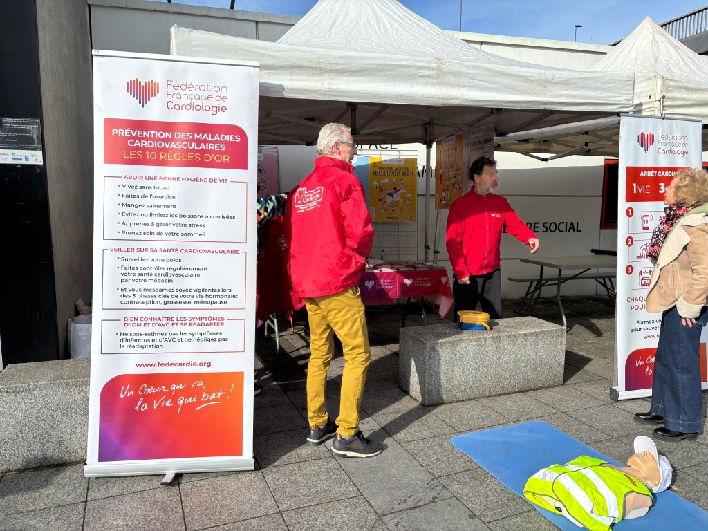 Stand de la Fédération Française de Cardiologie avec des bénévoles en vêtements rouges, mettant en avant la prévention des maladies cardiovasculaires. Une démonstration de réanimation cardio-respiratoire est en cours, avec un mannequin sur le sol et des affiches éducatives en arrière-plan.