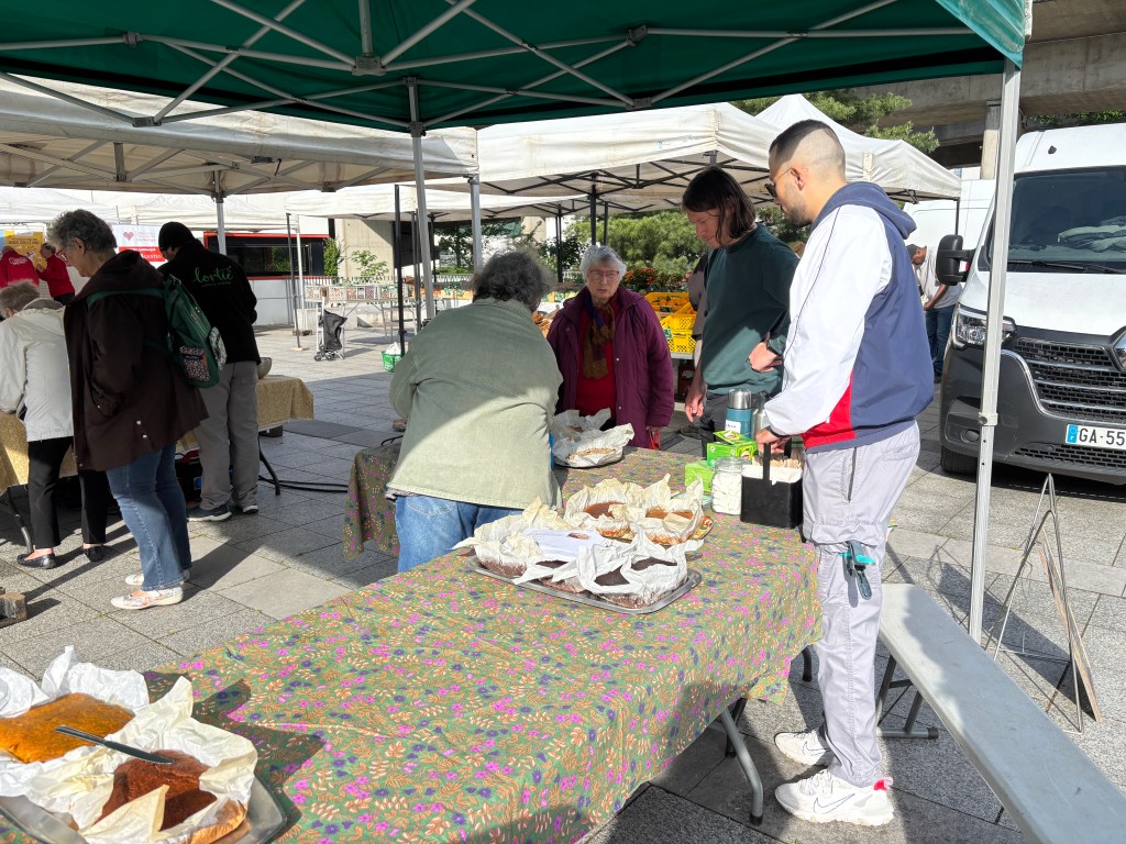 Un marché en plein air avec des personnes autour d'une table chargée de produits alimentaires. Des tentes blanches sont installées, et des clients discutent et choisissent des produits.