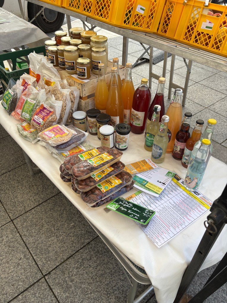 Table de marché avec divers produits alimentaires, y compris des confitures, des boissons artisanales, des pâtes, des légumineuses et des dattes, entourée de paniers en plastique orange.