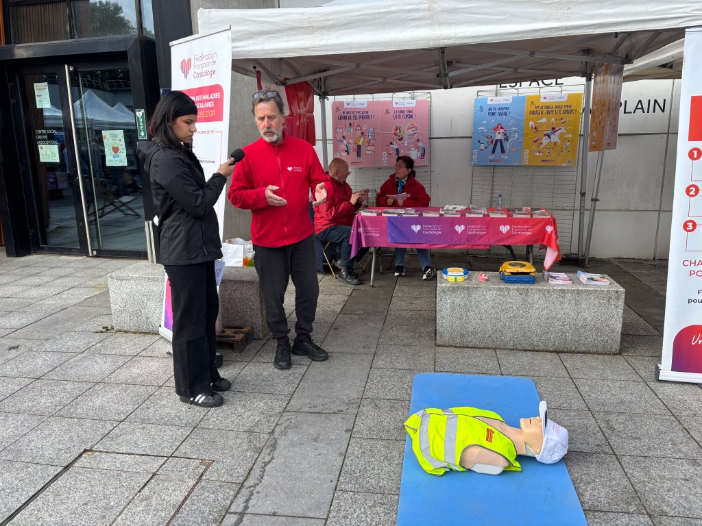 Un stand d'information sur la santé cardiaque avec des bénévoles, une personne interviewant un homme en gilet rouge, et un mannequin de réanimation sur un tapis bleu.