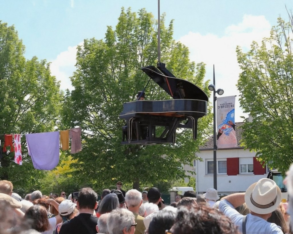 Un piano noir suspendu au-dessus d'une foule lors d'un événement en plein air, avec des arbres en arrière-plan et des vêtements accrochés à une corde à linge.