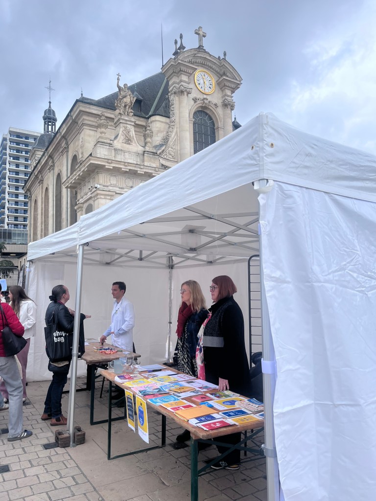 Un stand avec des personnes discutant devant une tente blanche, proche d'un bâtiment historique avec une horloge, sous un ciel nuageux.