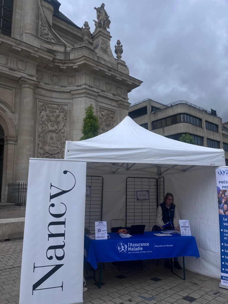 Un stand de l'Assurance Maladie à Nancy, avec une tente blanche et une femme en train de travailler à l'intérieur.