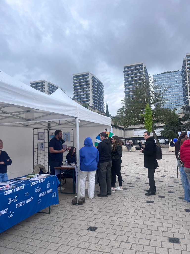 Des étudiants se rassemblent autour d'un stand d'information sous un auvent, avec des bâtiments modernes en arrière-plan et un ciel nuageux.