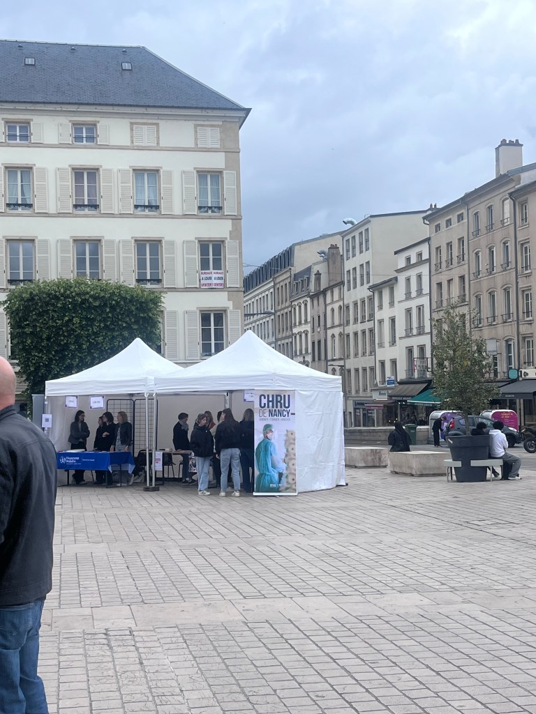 Un stand blanc dans une place de ville avec des personnes interagissant à l'intérieur, des bâtiments historiques en arrière-plan et un ciel nuageux.