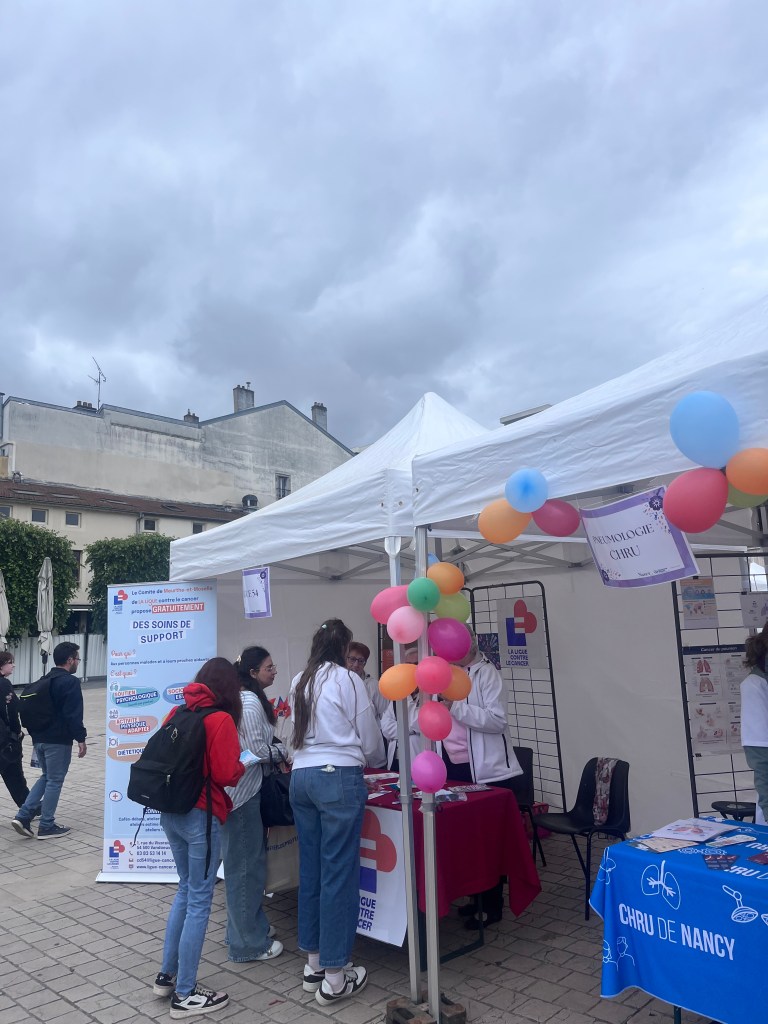 Un stand d'information avec des bénévoles portant des t-shirts blancs et des décorations colorées, lors d'un événement extérieur. Des affiches portent des informations sur le soutien aux personnes atteintes de cancer.