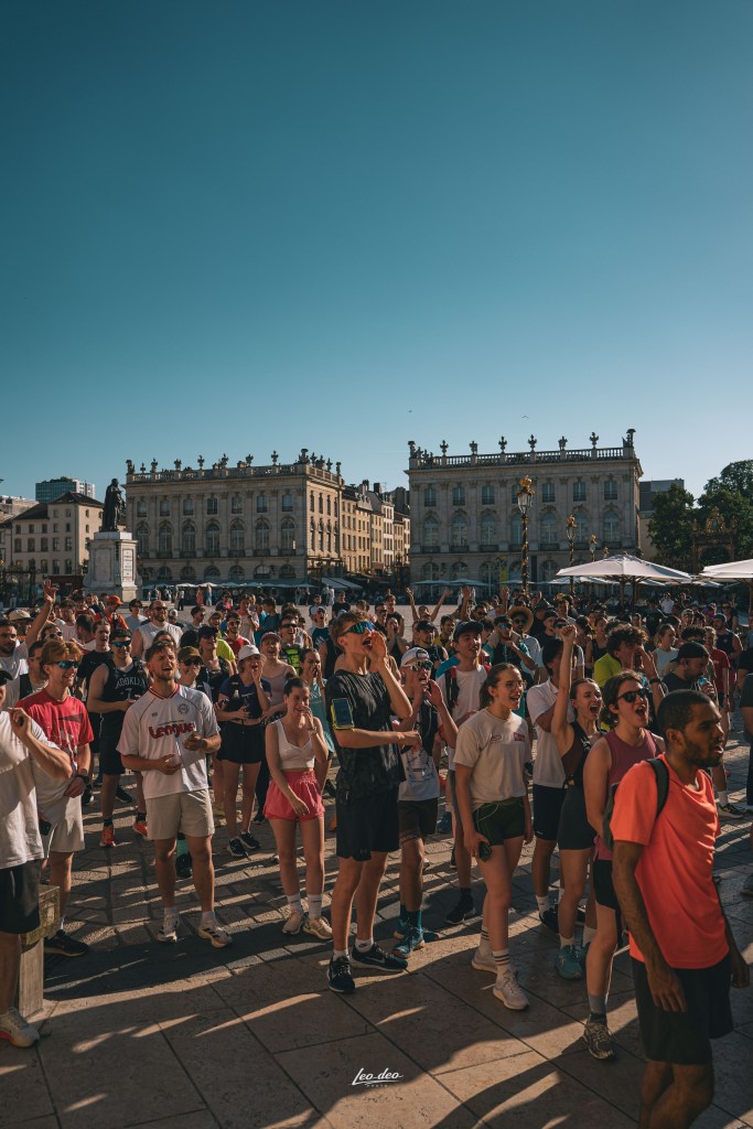 Une foule enthousiaste de jeunes adultes se rassemble sur une place ensoleillée, célébrant un événement ou un festival. Des bâtiments historiques et un ciel bleu clair sont visibles en arrière-plan.