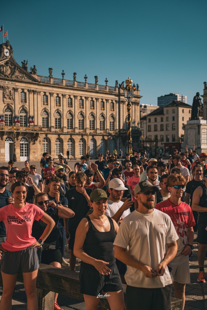 Un groupe de coureurs rassemblés sur une place ensoleillée, avec un bâtiment historique en arrière-plan.