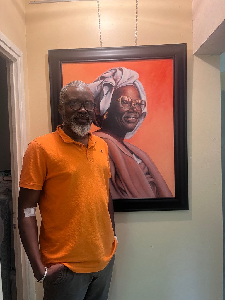 Un homme en polo orange se tient devant un tableau représentant un portrait d'une femme souriante portant un foulard, dans un intérieur lumineux.