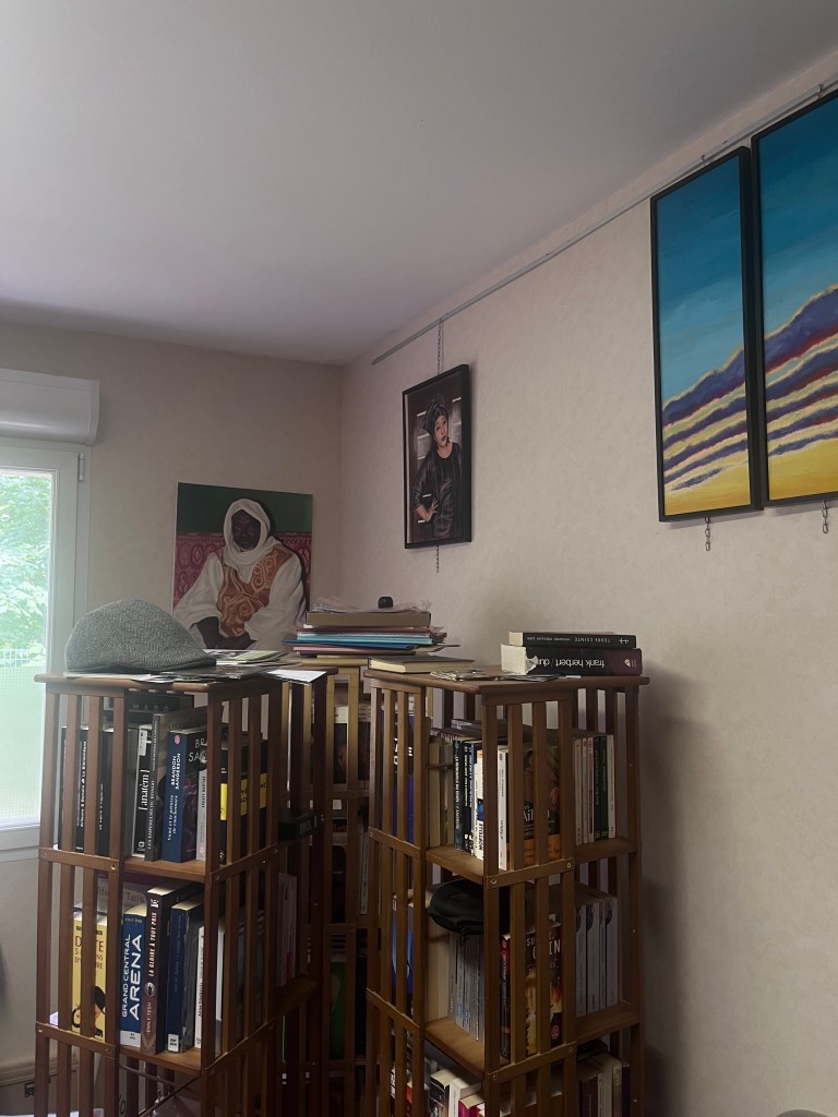 Une bibliothèque en bois avec des livres sur plusieurs étagères, un chapeau sur le dessus. Au mur, deux tableaux avec des paysages et un portrait d'une personne.