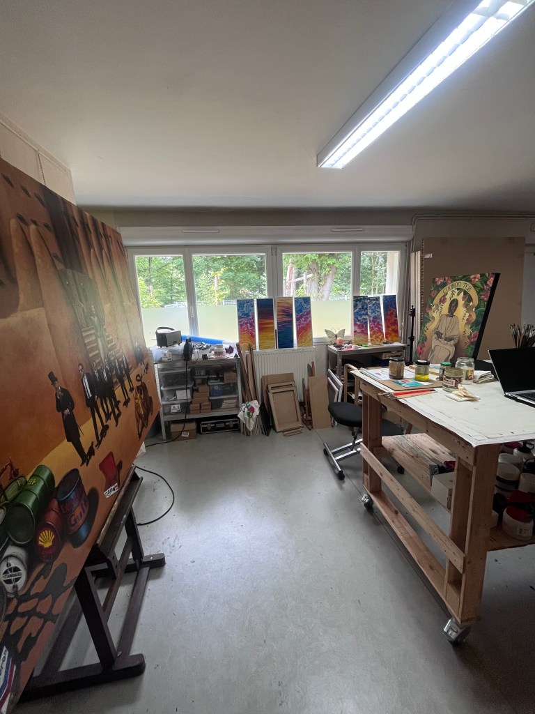 Un atelier d'art avec un grand tableau sur le mur gauche, des peintures colorées exposées à la lumière du jour à droite, et une table de travail en bois avec des fournitures artistiques.