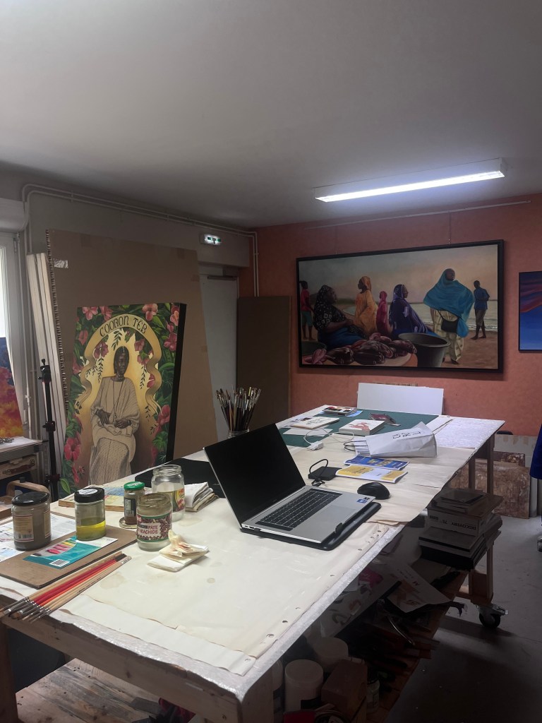 Un atelier d'artiste avec un bureau encombré de fournitures telles que des pinceaux, de la peinture et un ordinateur portable. Des œuvres d'art, y compris une grande toile accrochée au mur et des affiches, sont visibles en arrière-plan.