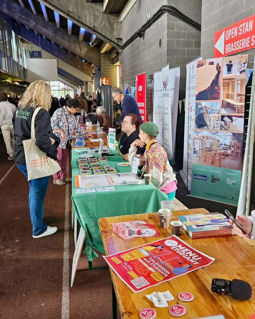 Un salon avec plusieurs stands, des tables occupées par des exposants et des visiteurs interagissant, sous un éclairage industriel et un environnement animé.